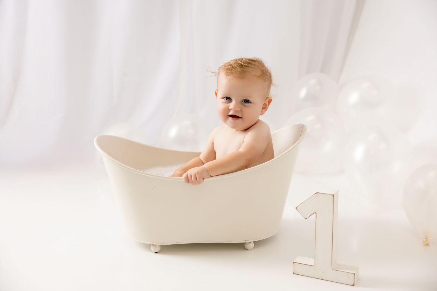 Smiling baby sitting in a white mini bathtub with balloons and a number 1 sign for a first birthday photo.
