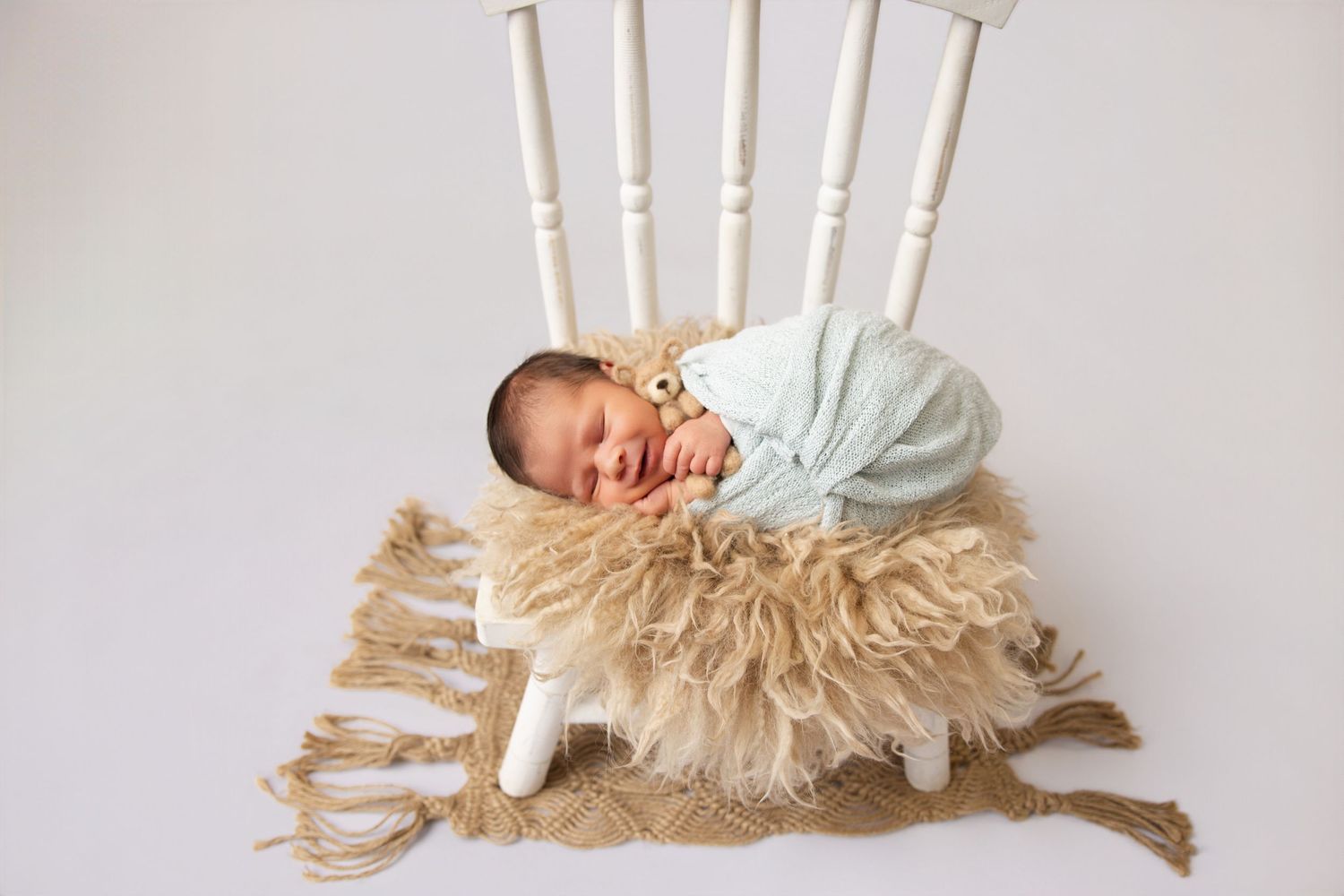 Swaddled newborn baby sleeping on a fluffy rug beneath a white wooden chair with a small teddy bear.