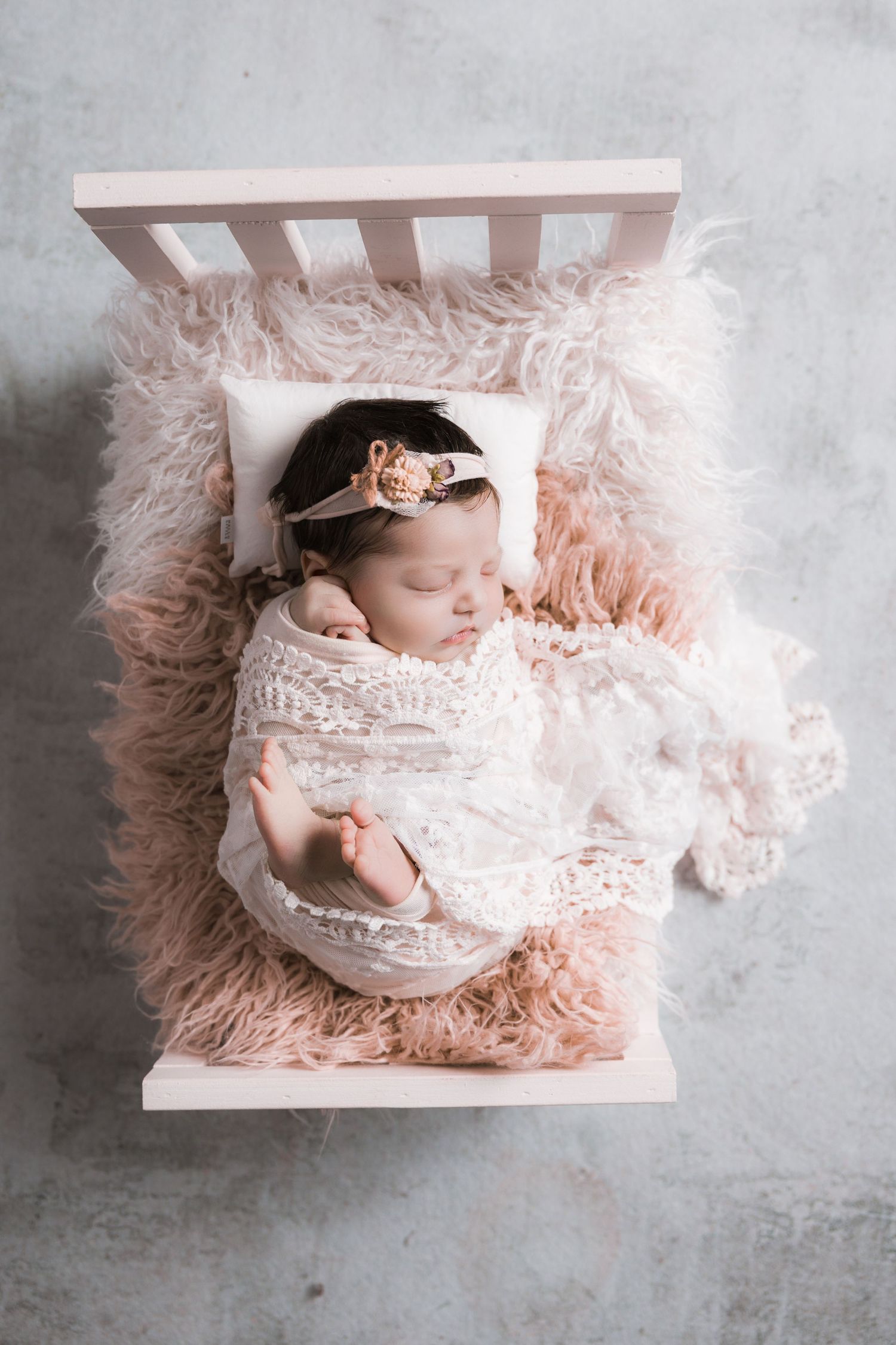 Newborn baby girl swaddled in white lace wrap, sleeping on pink fluffy fur in a miniature white crib.