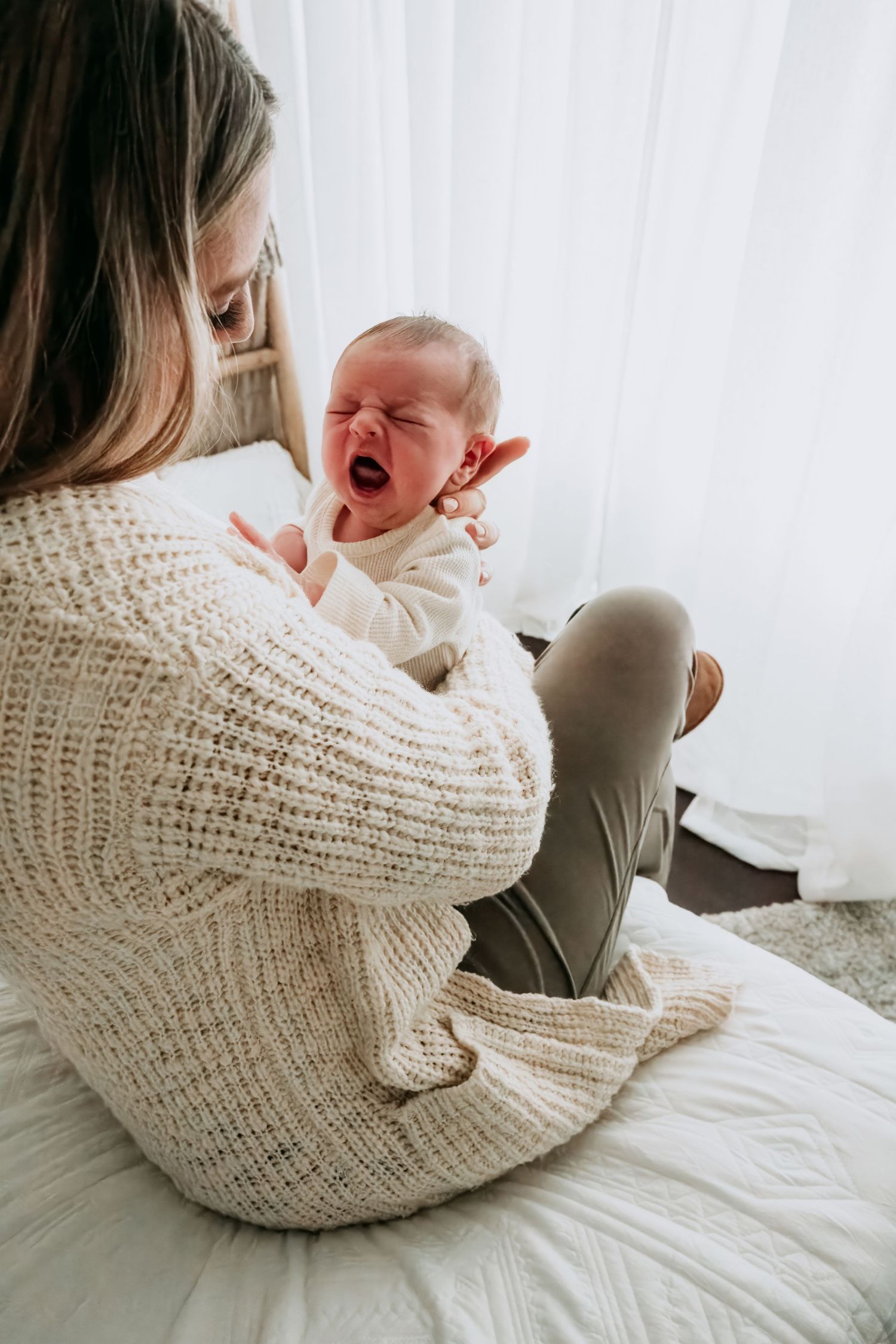 A mother in a cream knit sweater holds her crying newborn baby while sitting on a white bed near a window.