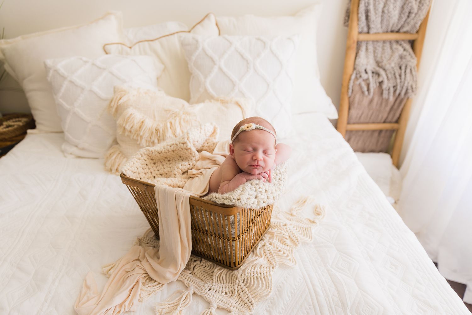 Newborn baby in cream wrap nestled in wicker basket on white bed in bright boho studio setting in the Bonsall Studio near Temecula