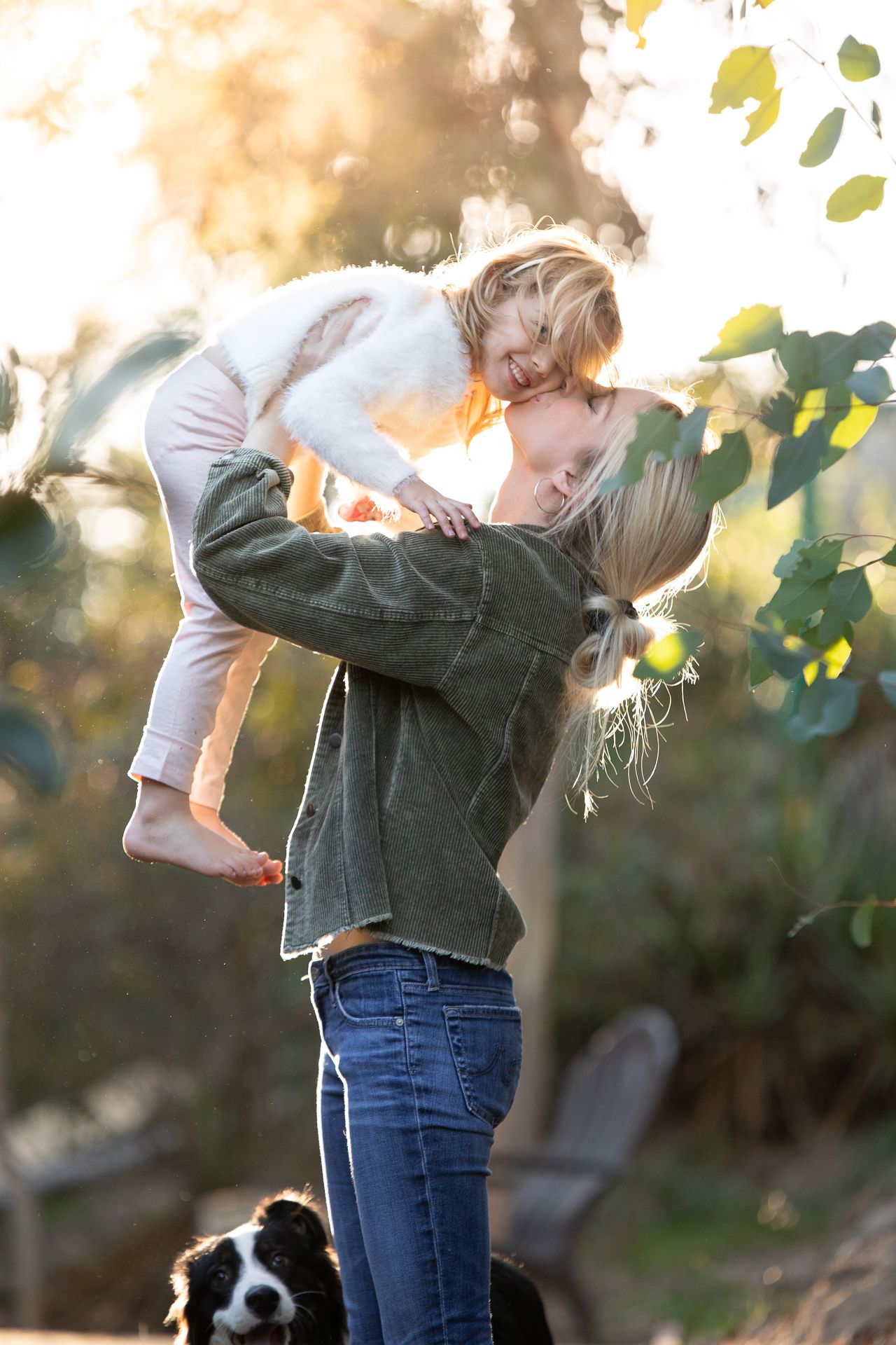 A blonde woman lifts a laughing child into the air outdoors with a dog nearby in golden light.