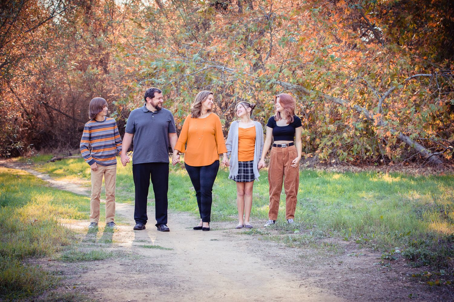 Family of five walks hand in hand on a dirt path surrounded by autumn foliage.