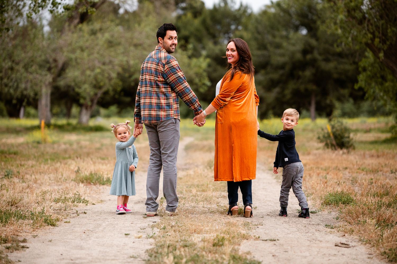 Pregnant family of four holding hands on a dirt path in a park.