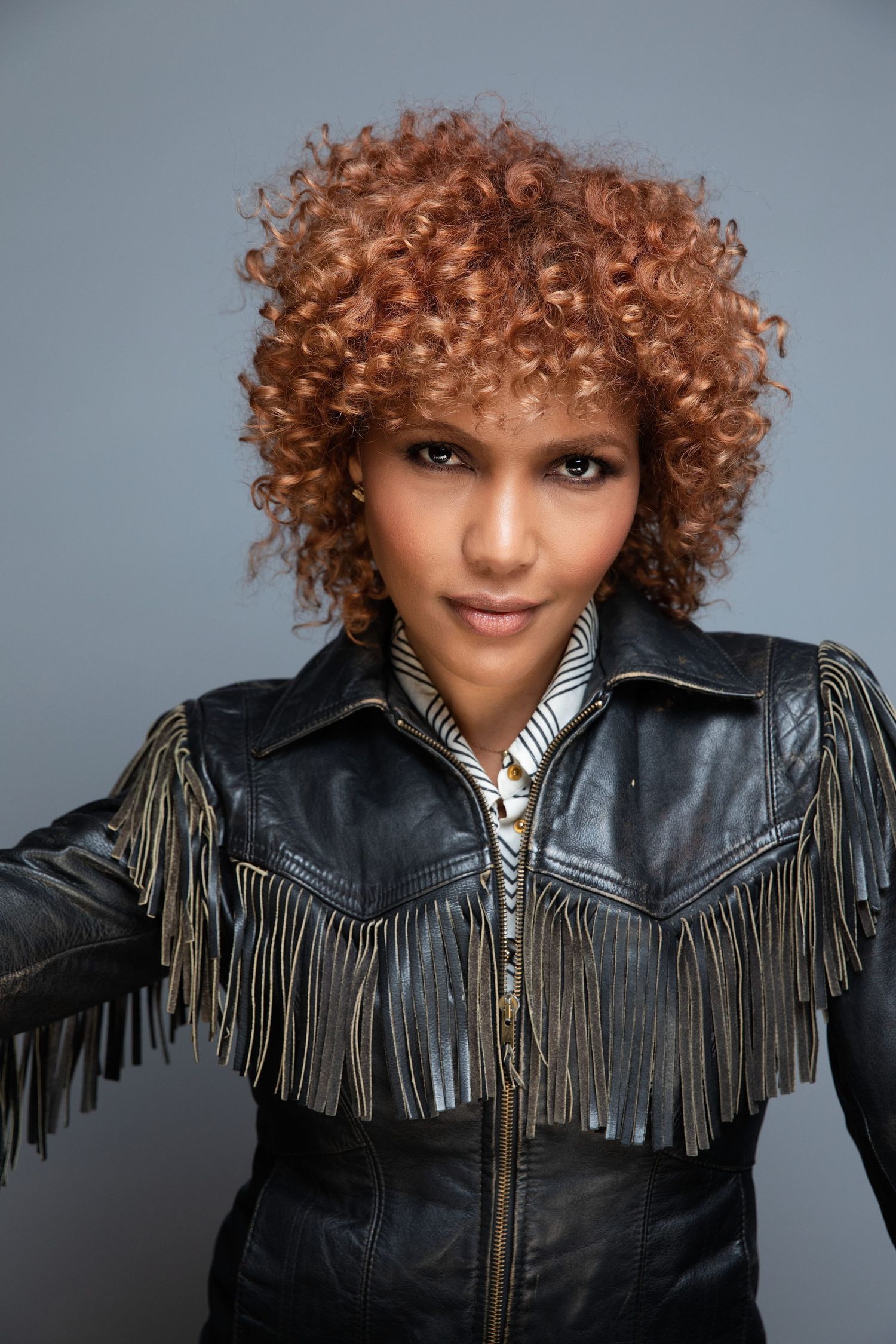 A personal branding session. Woman wearing a black leather fringe jacket poses against a gray backdrop in a dynamic photo series.