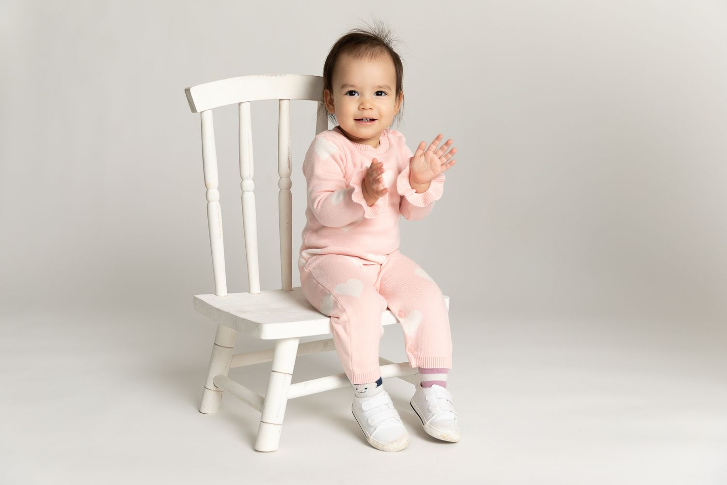 Happy baby in pink outfit clapping hands on white chair.