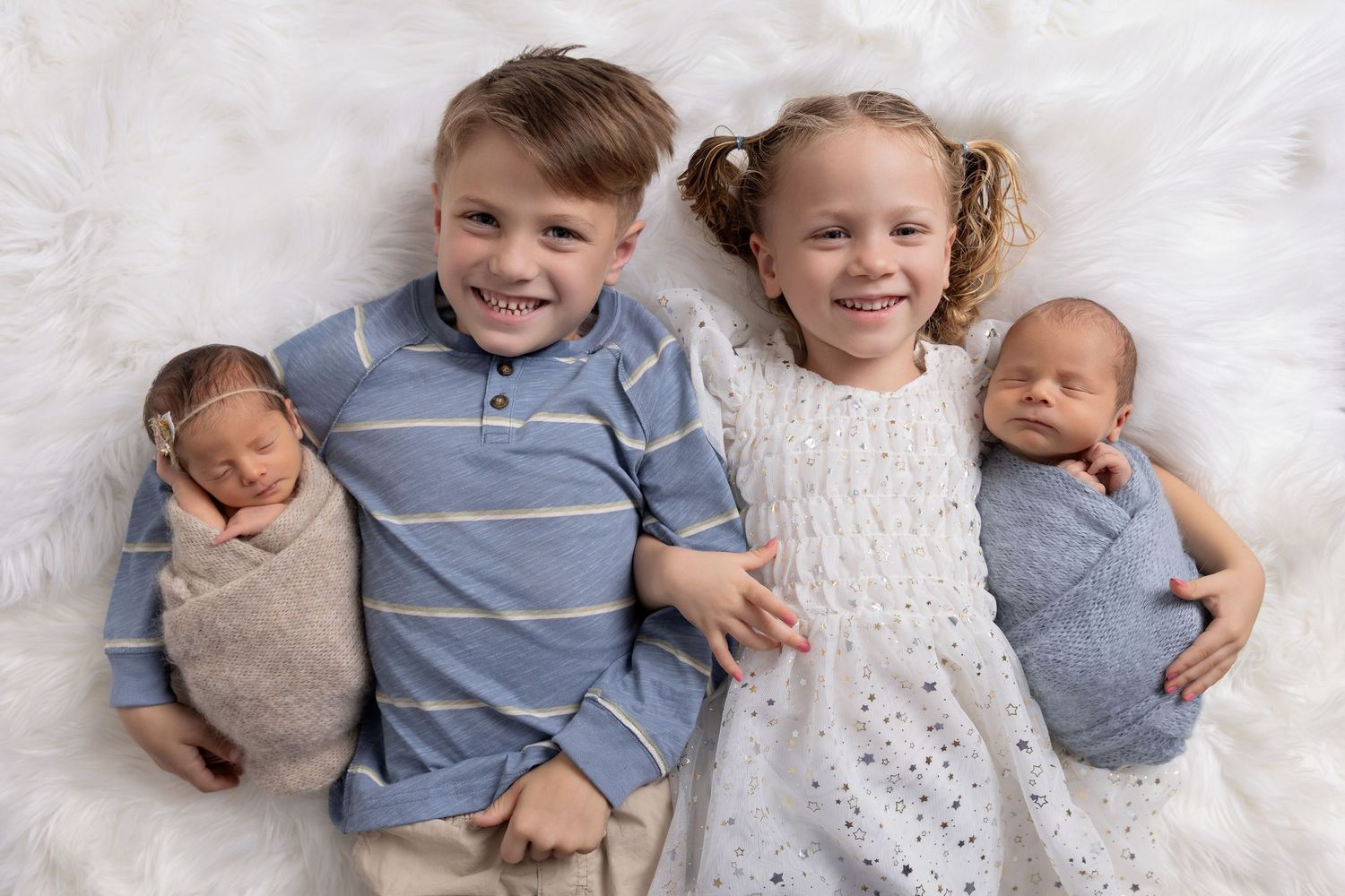 Two smiling children lying on white fur, each holding a swaddled newborn twin baby in a sweet family portrait.