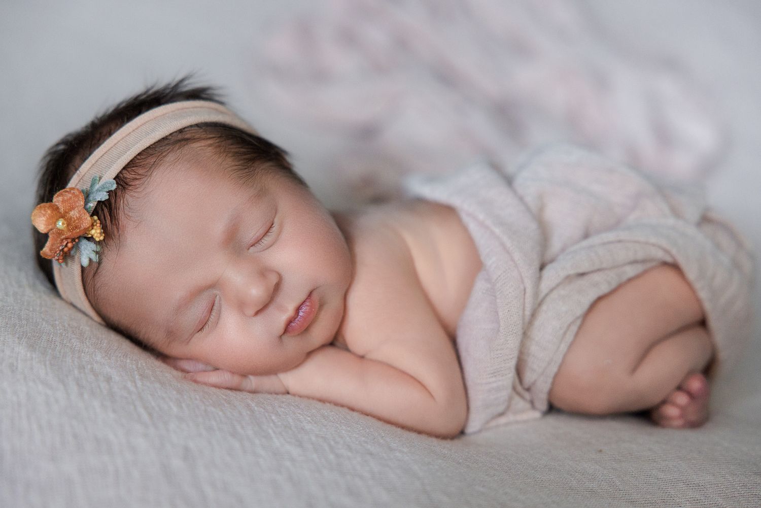 Peaceful newborn baby girl sleeping wrapped in soft blush fabric with a delicate floral headband.