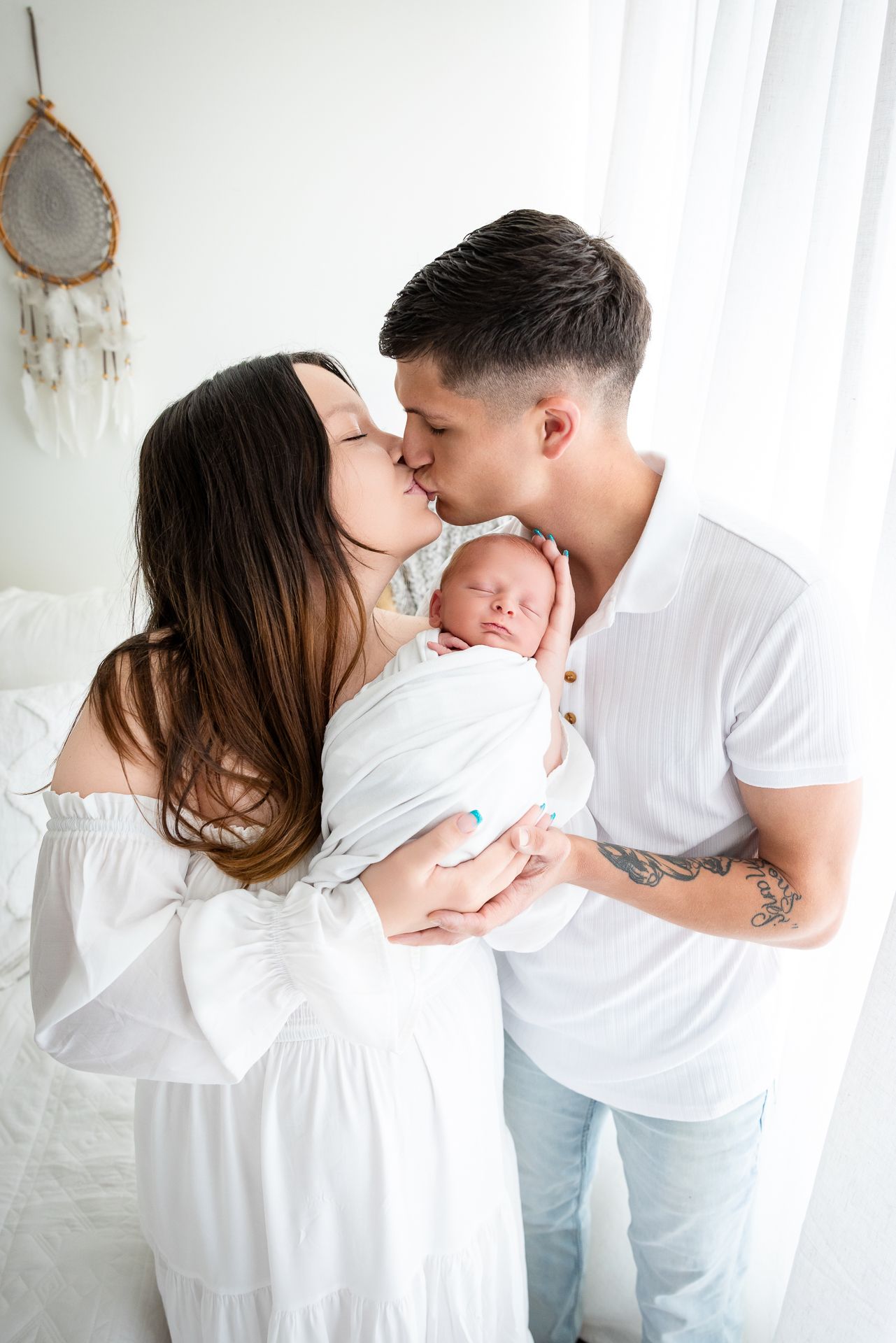 Young couple in white clothing kissing while tenderly holding their swaddled newborn baby in a bright, airy room.