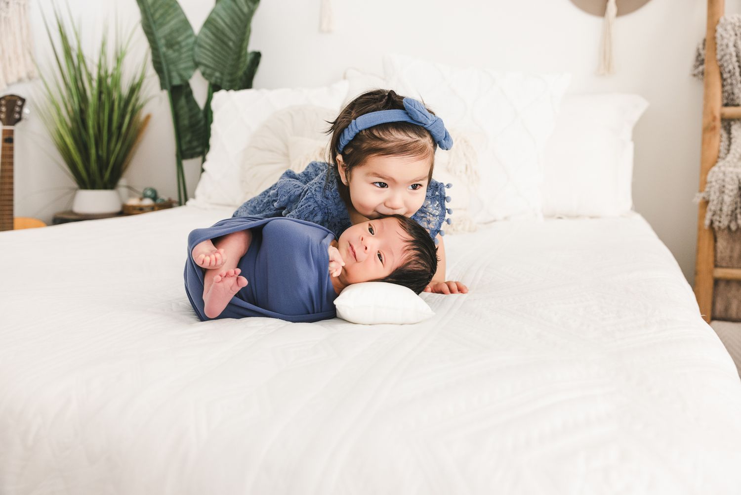 Toddler girl in denim dress hugging swaddled newborn baby sibling on white bed with boho nursery decor.
