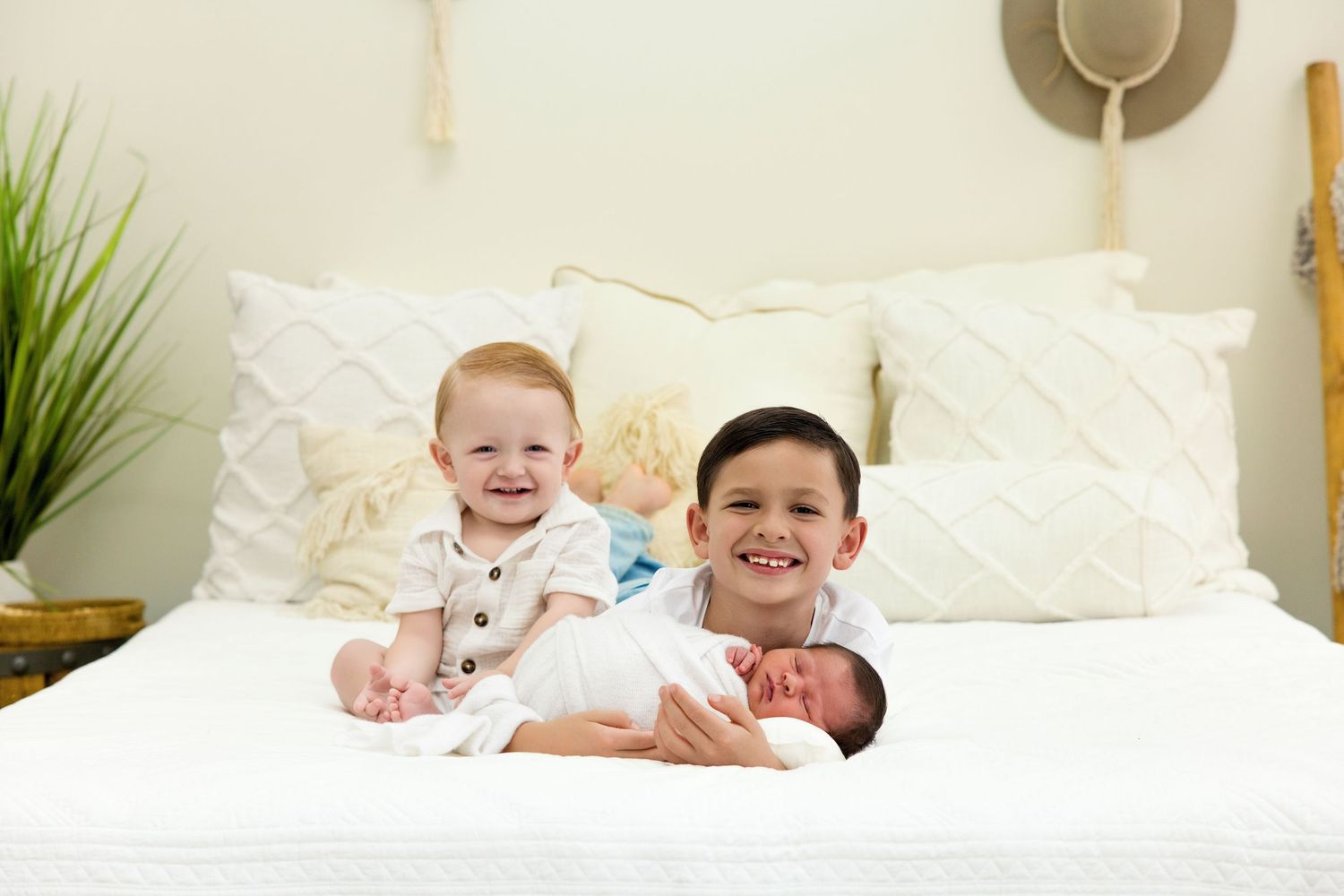 Two smiling boys sit on a white bed holding their newborn sibling, surrounded by fluffy pillows.