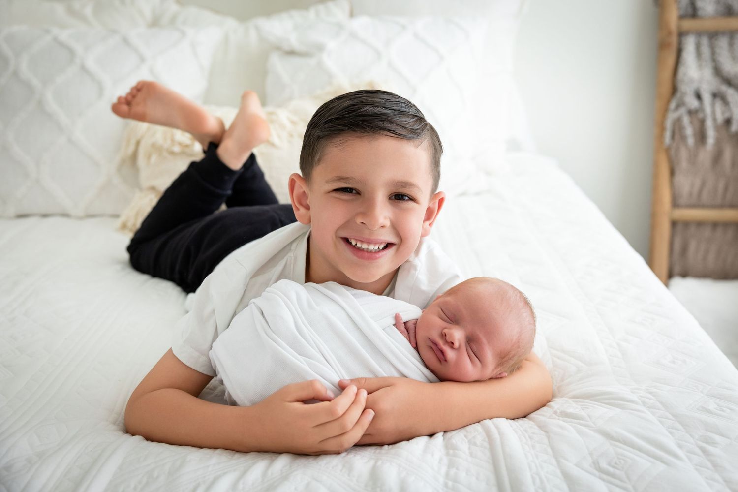 Smiling boy lying on white bed gently holding swaddled newborn baby sibling in bright, cozy room.