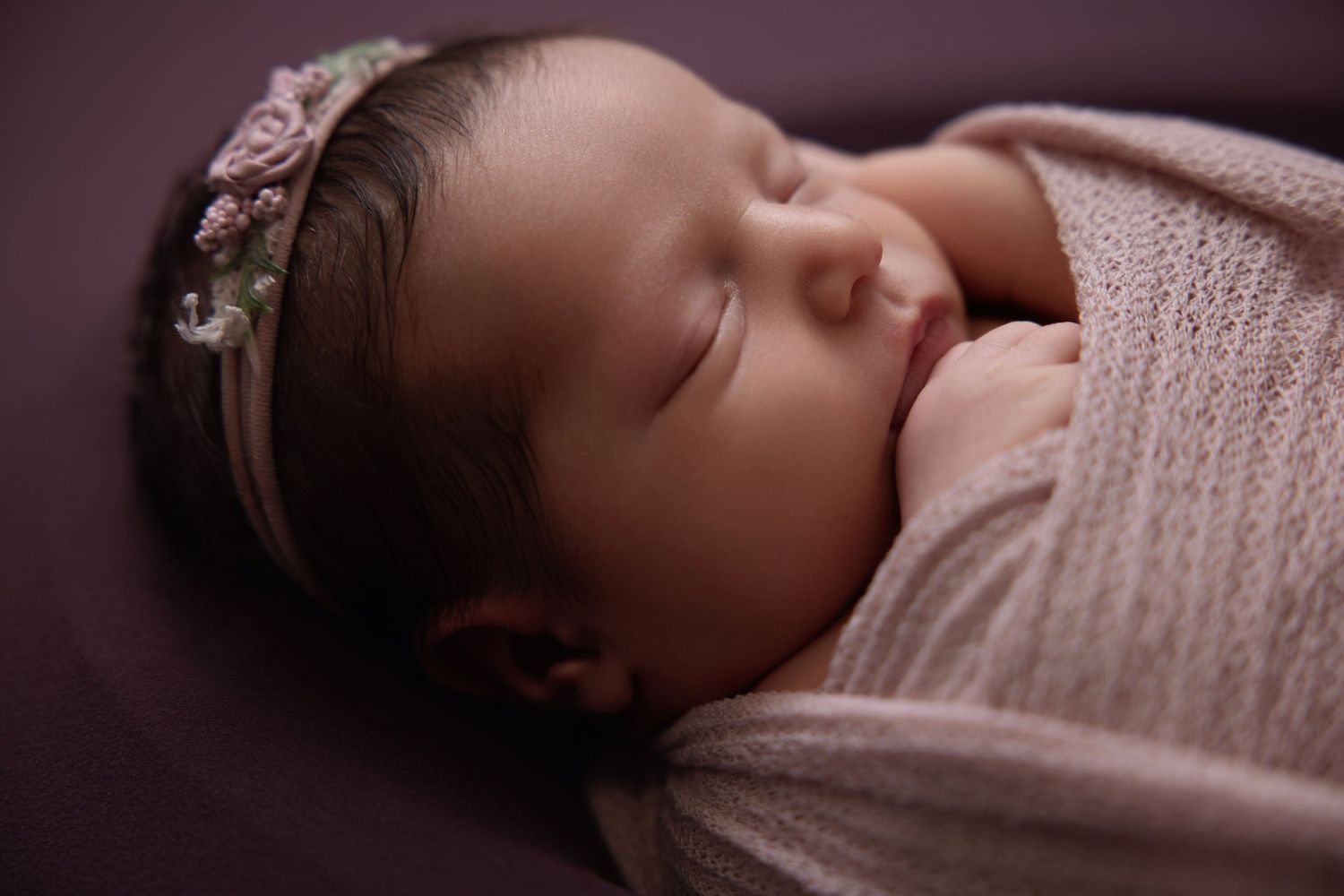 Sleeping newborn baby girl wrapped in pink swaddle with floral headband on purple background.