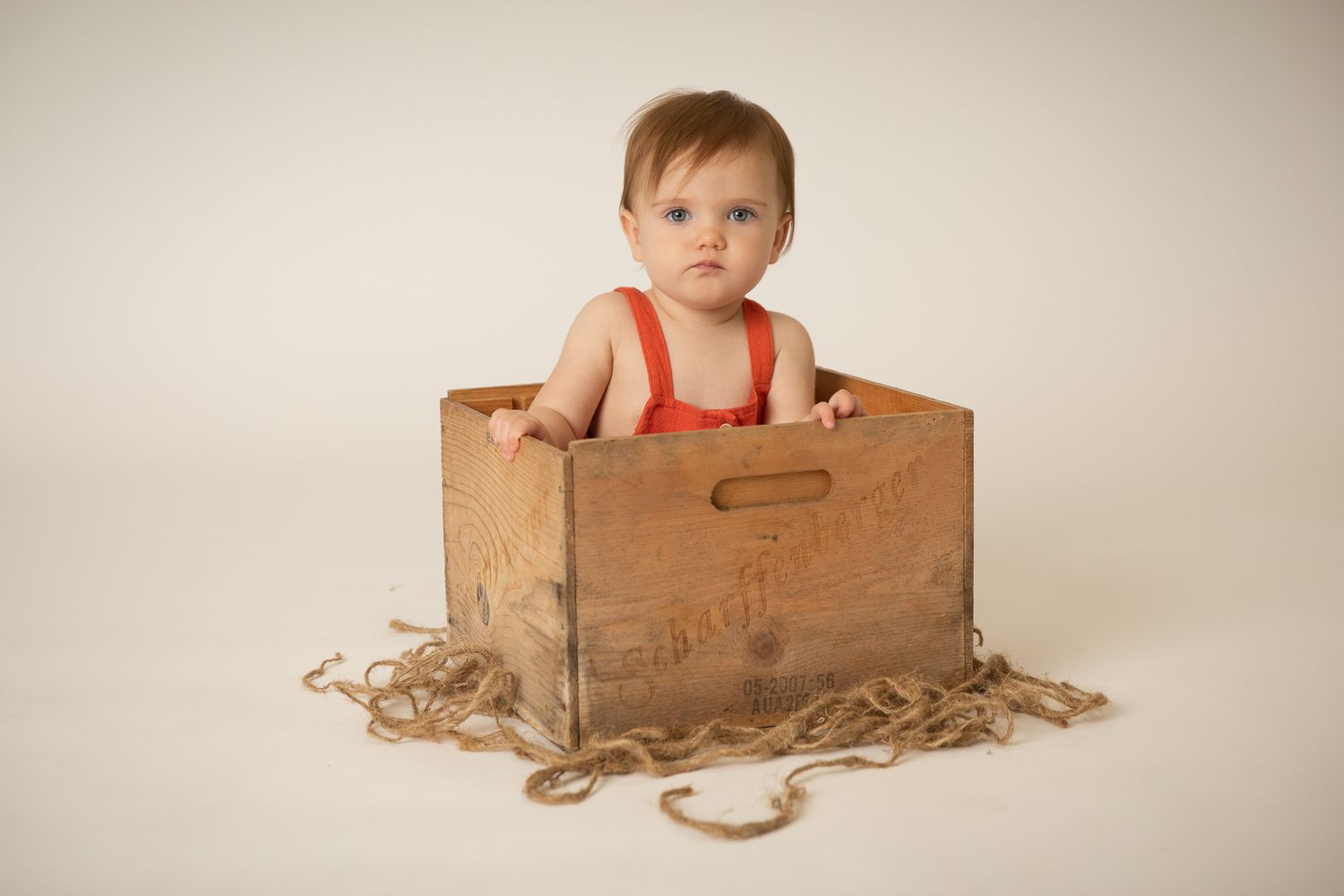 Baby in red romper sitting inside a rustic wooden crate on a white background.