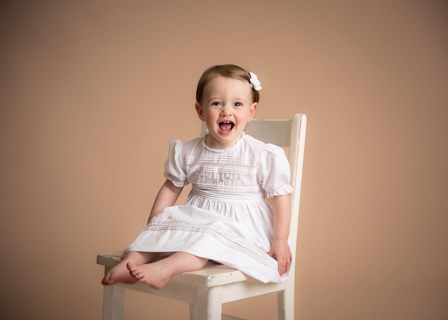 Laughing toddler girl in white smocked dress sitting on white chair, beige backdrop.