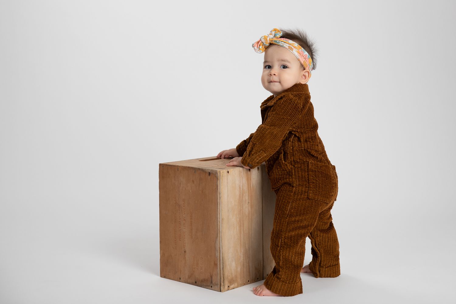 Baby girl in brown corduroy romper standing beside rustic wooden crate.