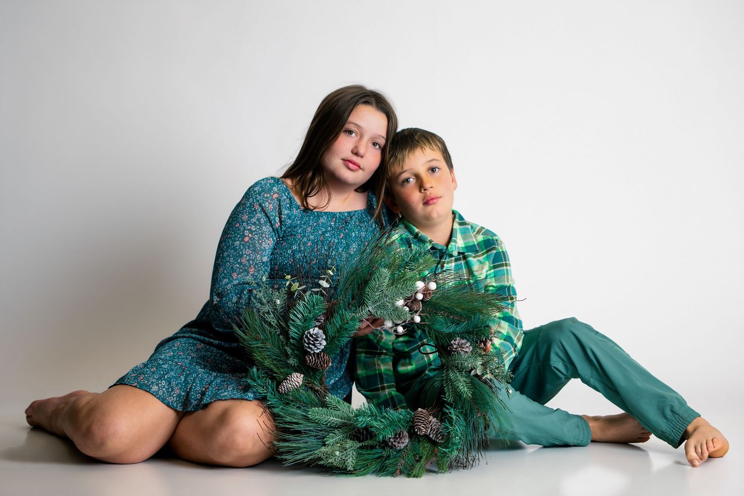A teenage girl and young boy sit together in teal outfits holding festive green pine branches in a studio.