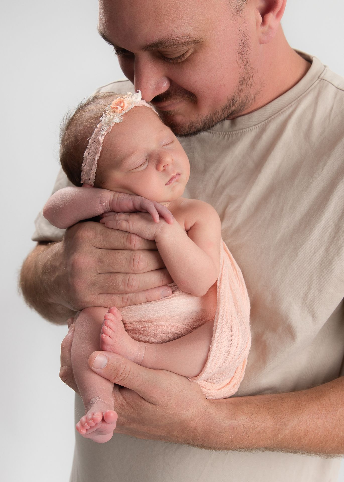 A father tenderly kisses his sleeping newborn baby girl wrapped in pink, wearing a floral headband.