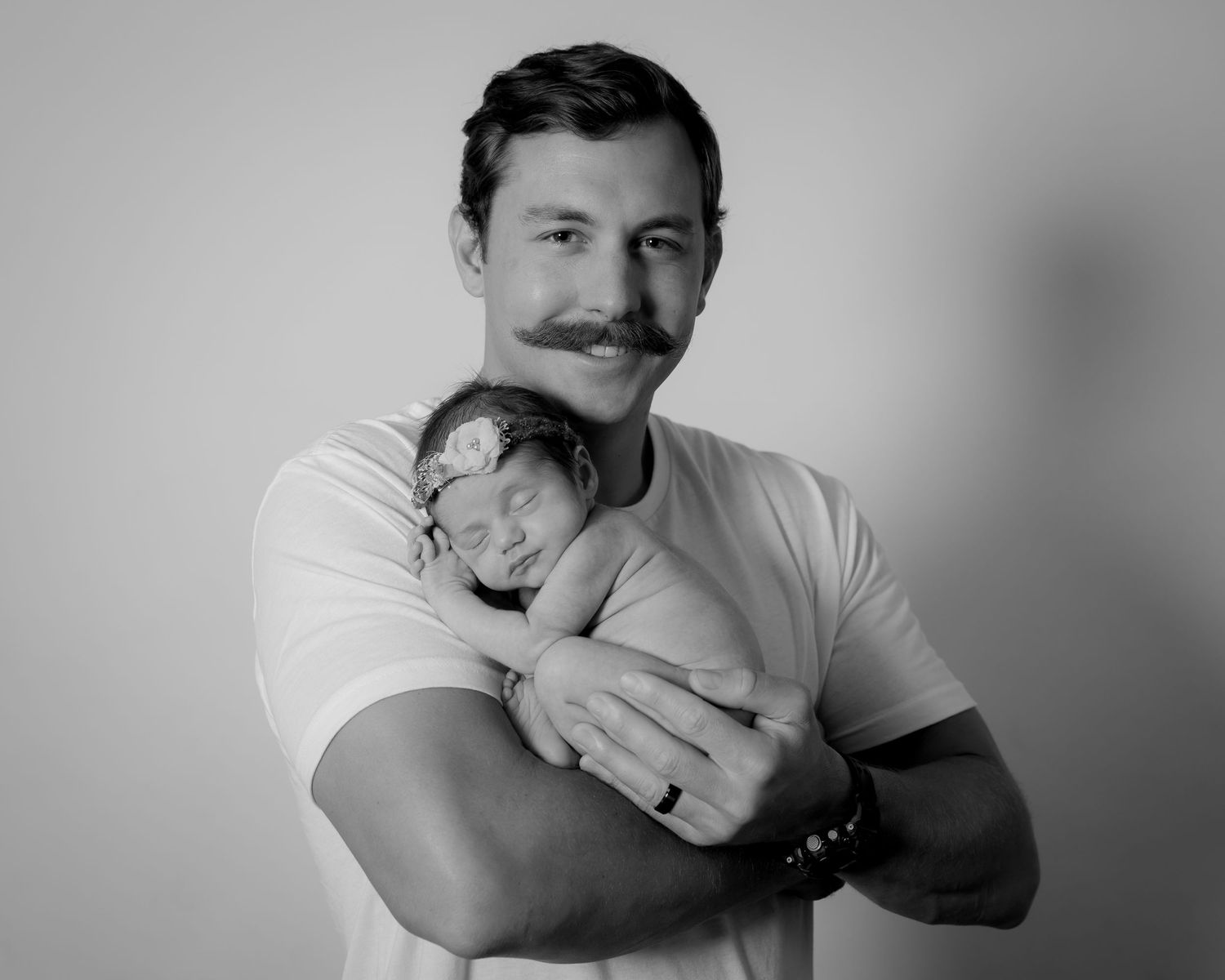 Black and white photo of a smiling man with mustache cradling a sleeping newborn baby against his chest.