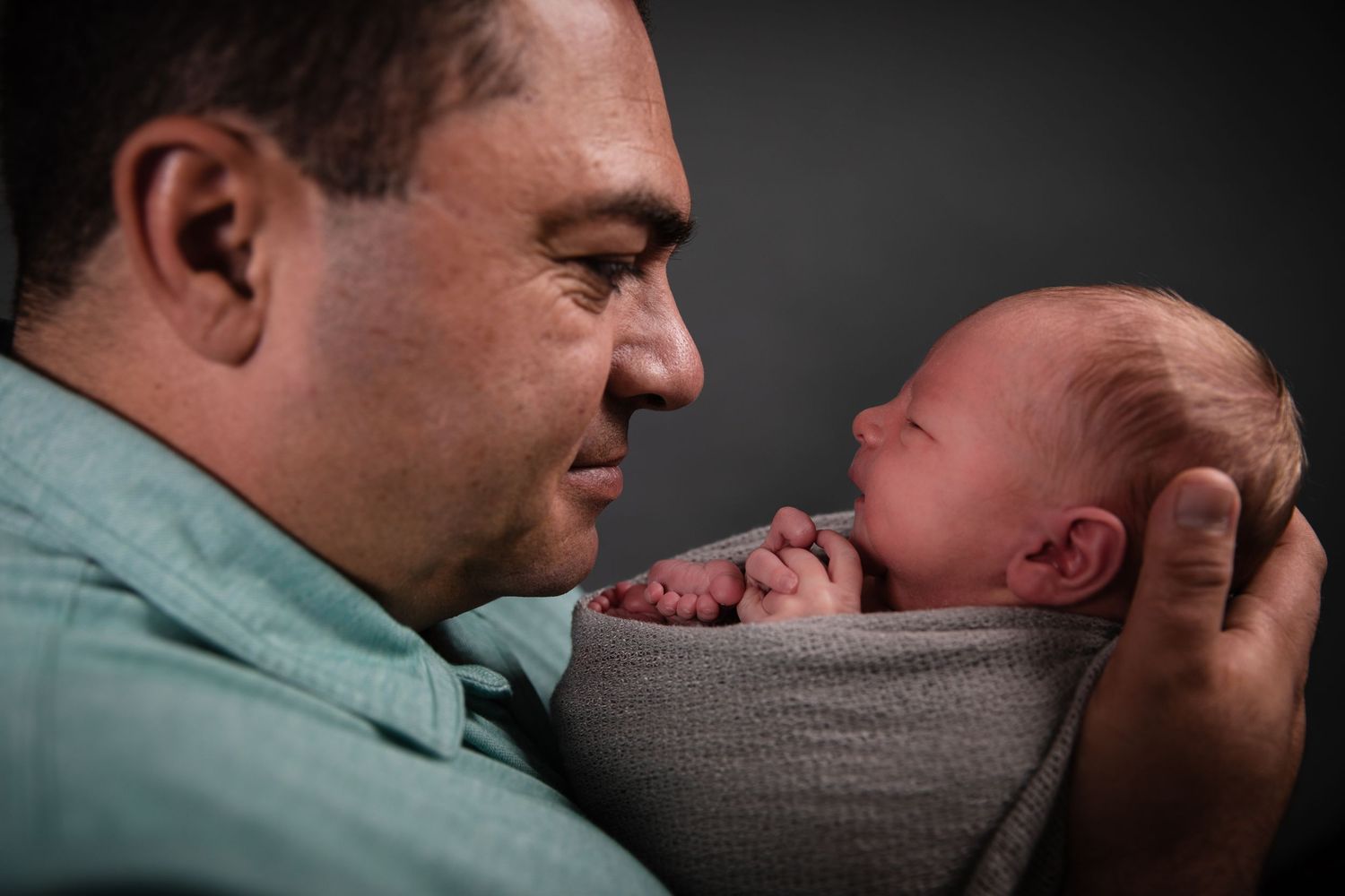 A father tenderly holds his swaddled newborn baby close, sharing a precious bonding moment against a dark background.