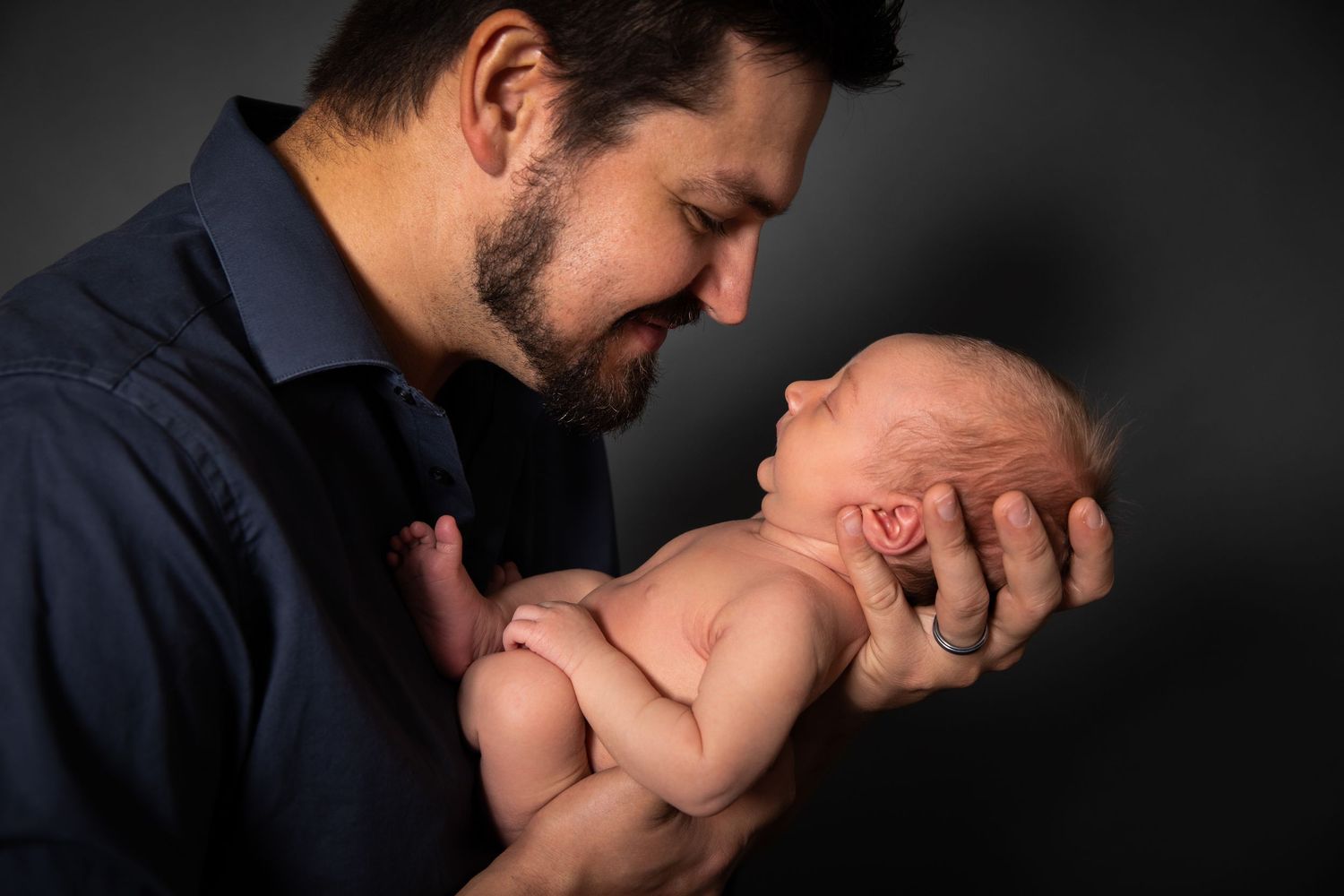 A bearded man in a blue shirt tenderly holds a sleeping newborn baby against a dark background.