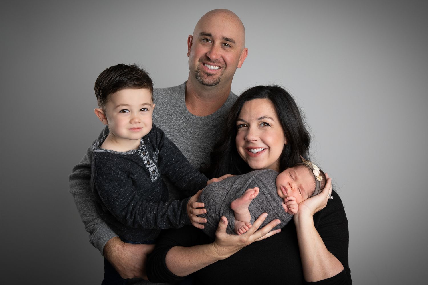 Family portrait with father holding toddler son, mother cradling newborn baby, smiling together against gray studio backdrop.