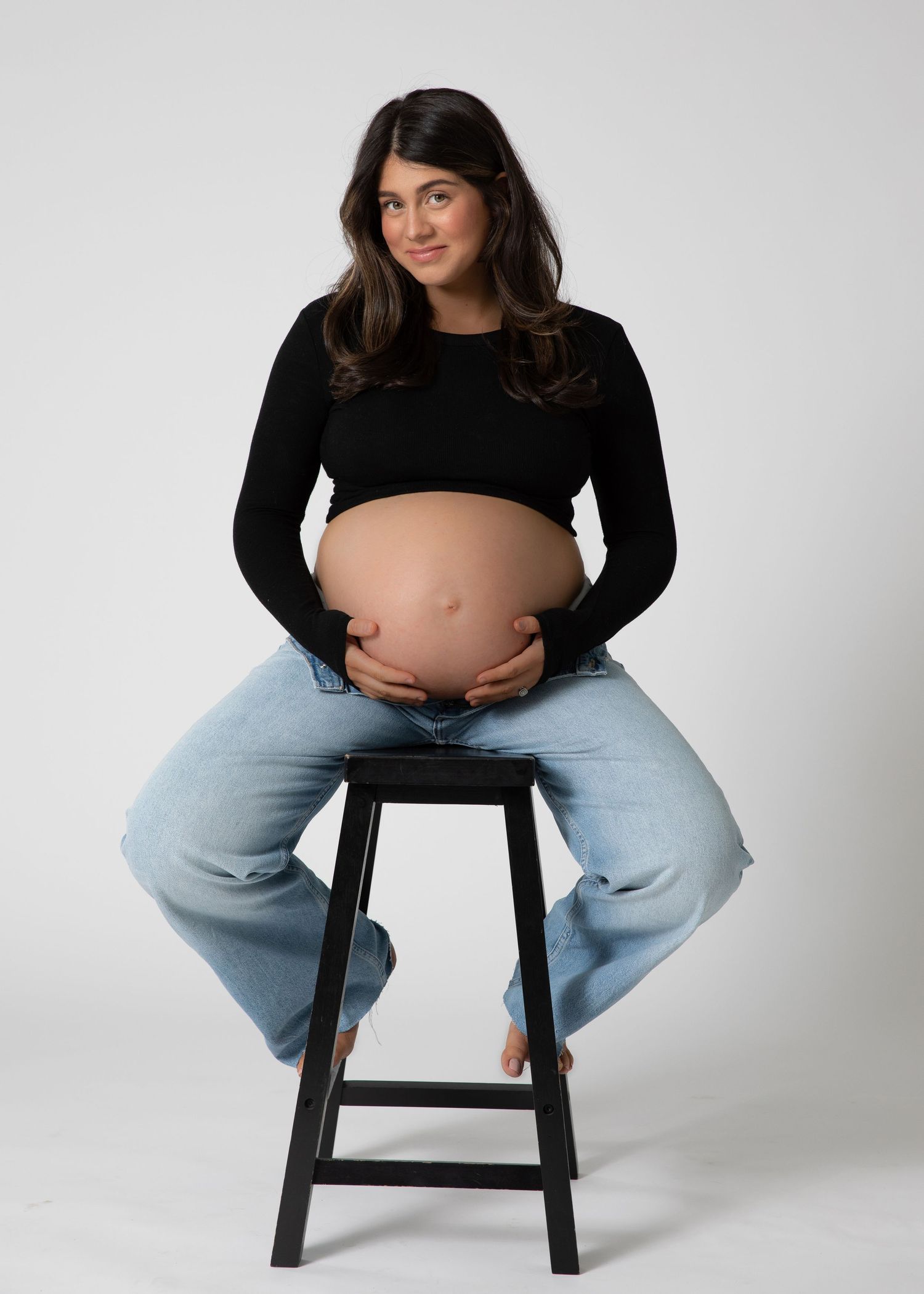 Pregnant brunette woman sitting on stool holding baby bump in black crop top in Bonsall, near Fallbrook, & Temecula