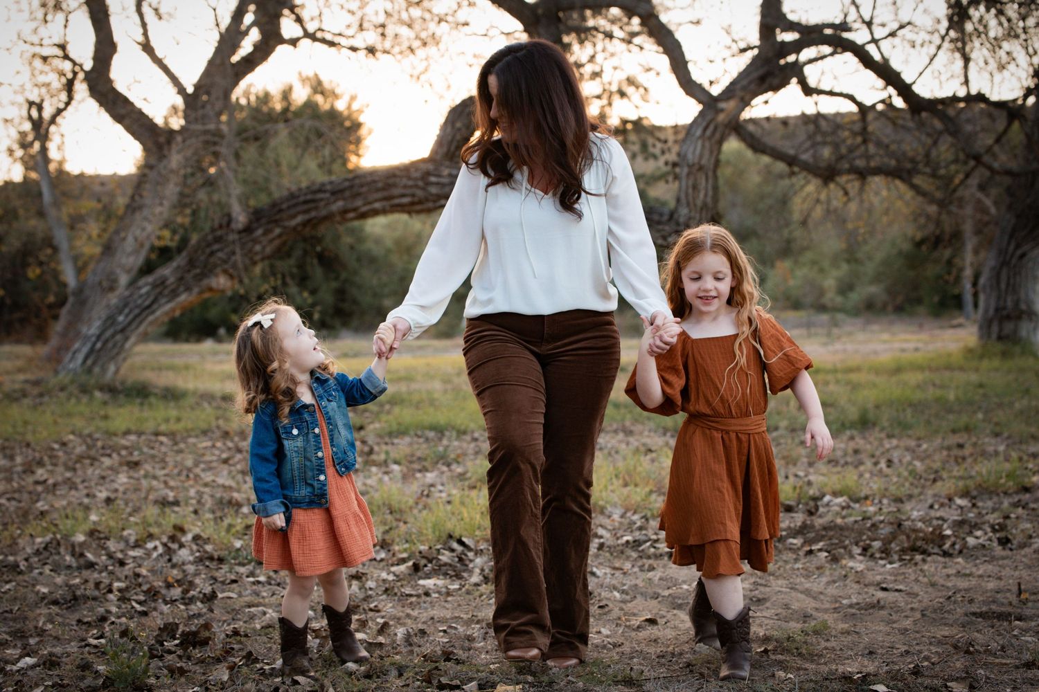 Mother walking hand-in-hand with two young daughters through a sunlit outdoor nature setting.