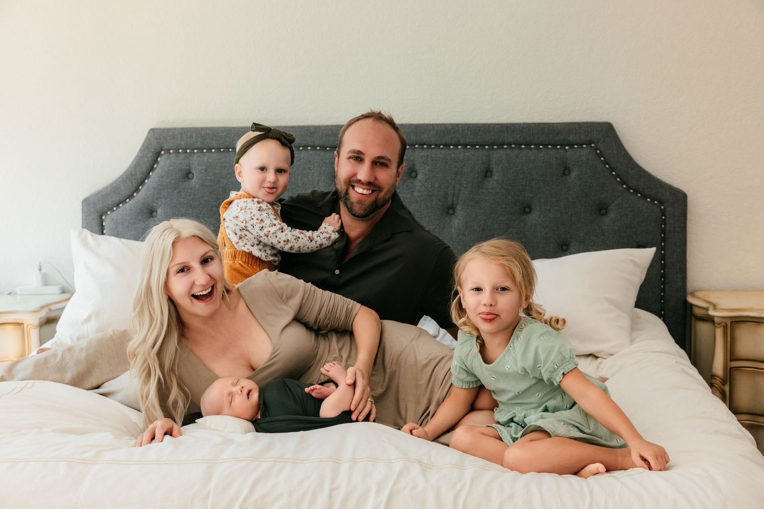 Happy family of four posing together on a white bed with two young daughters and a newborn baby.
