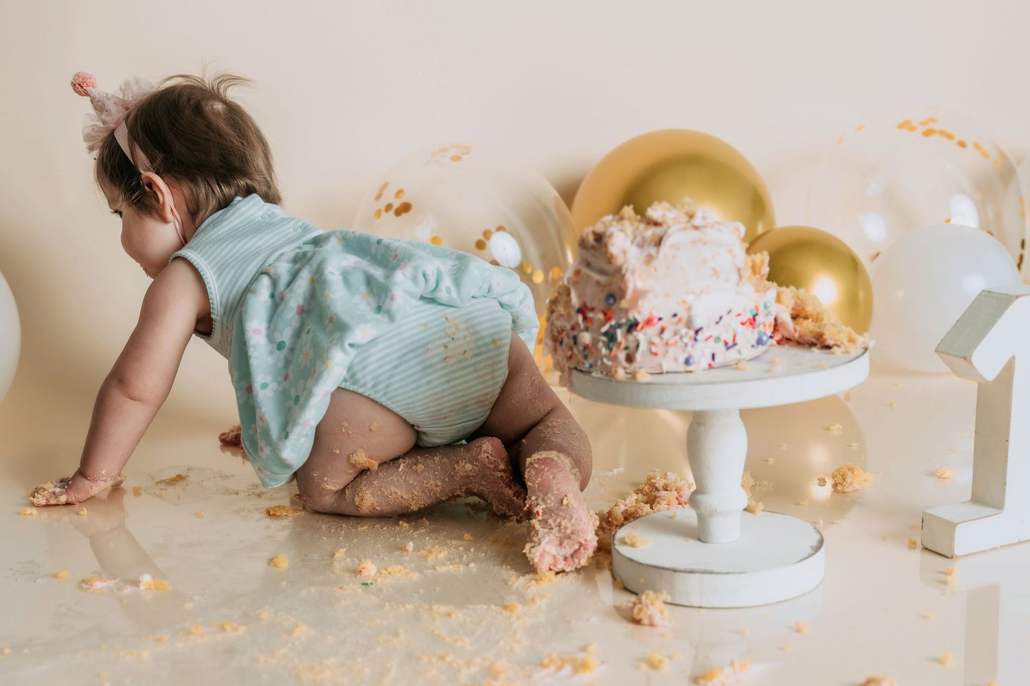 First Birthday Session. Baby in teal romper crawling away from smashed birthday cake on white floor in Fallbrook Newborn studio