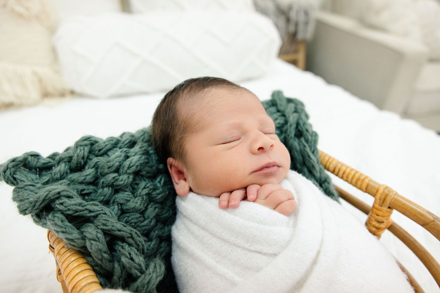 Sleeping newborn baby swaddled in white wrap resting in wicker basket with green chunky knit blanket.