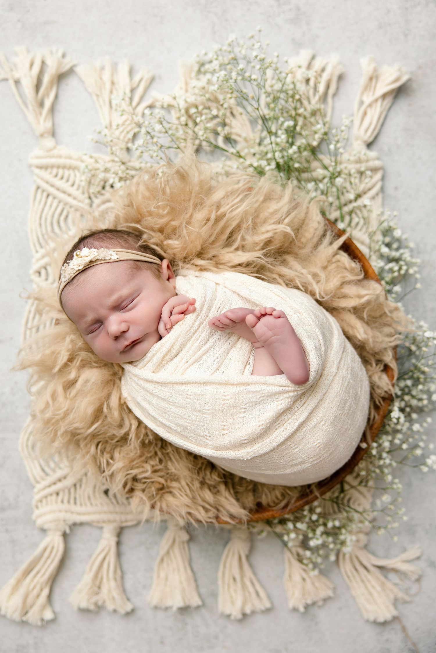 Sleeping newborn baby swaddled in cream wrap, resting in a basket with fluffy fur and baby's breath flowers on macrame mat.