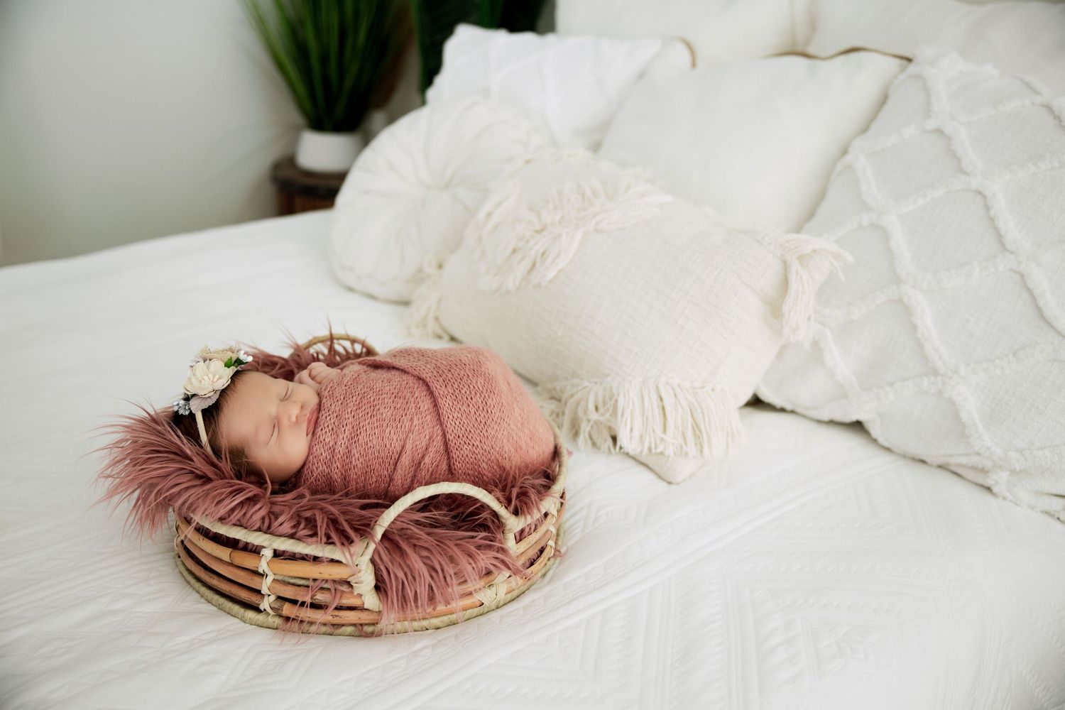 Newborn baby swaddled in pink wrap, sleeping in a wicker basket with fur lining on a white bed.