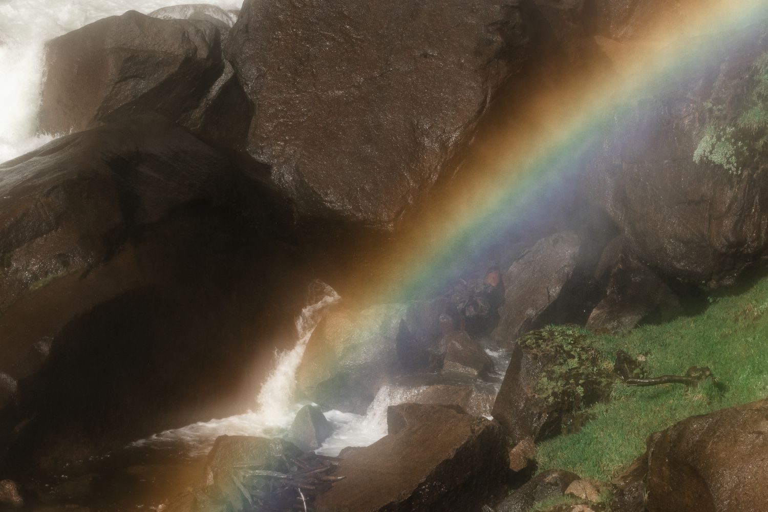 A vibrant rainbow arcs over rushing waterfall cascading through dark rocky gorge surrounded by lush green moss.