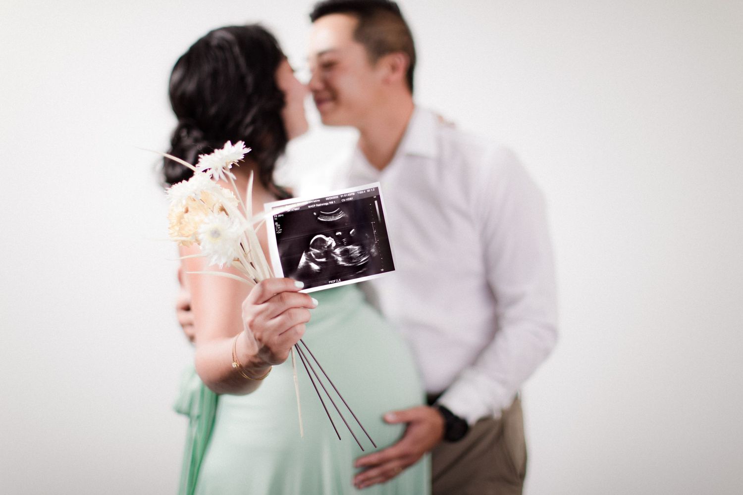Couple kissing while pregnant woman holds ultrasound photo and decorative sticks in front of them in Bonsall, near Fallbrook, & Temecula