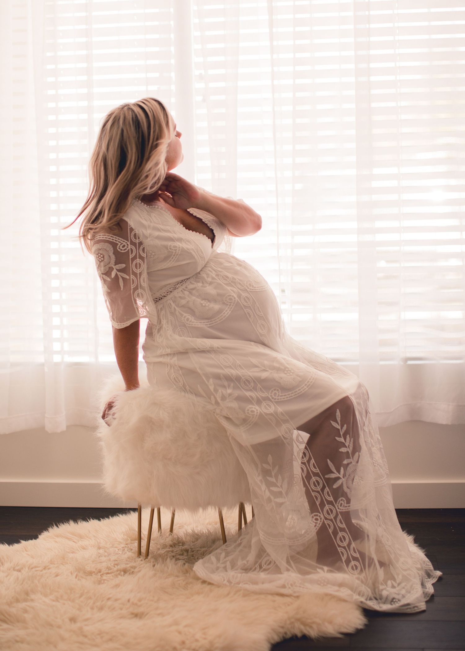 Pregnant woman in a white lace gown sitting gracefully on a fur stool near bright window blinds in Bonsall, near Fallbrook, & Temecula