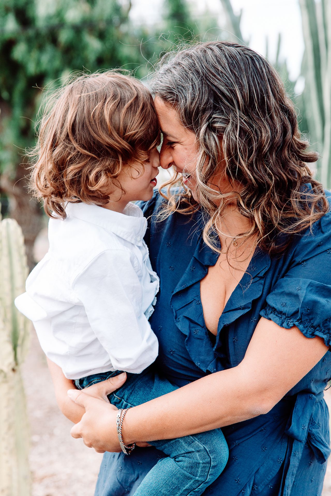 Mother and curly-haired toddler touching foreheads lovingly outdoors near green cactus plants in Bonsall, near Fallbrook, & Temecula