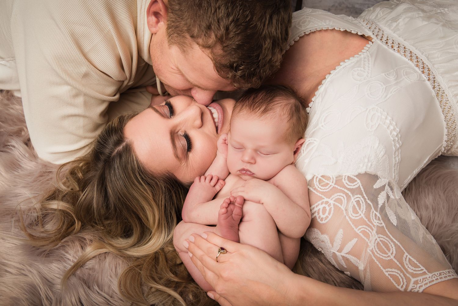 New parents tenderly kiss their sleeping newborn baby while lying together on a soft fur rug in Vista