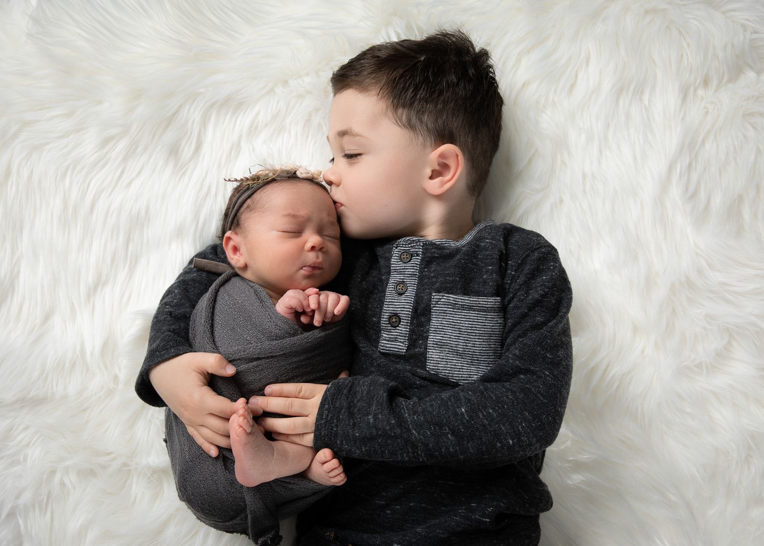 Older brother gently kissing newborn baby sibling while lying on white fluffy fur backdrop.