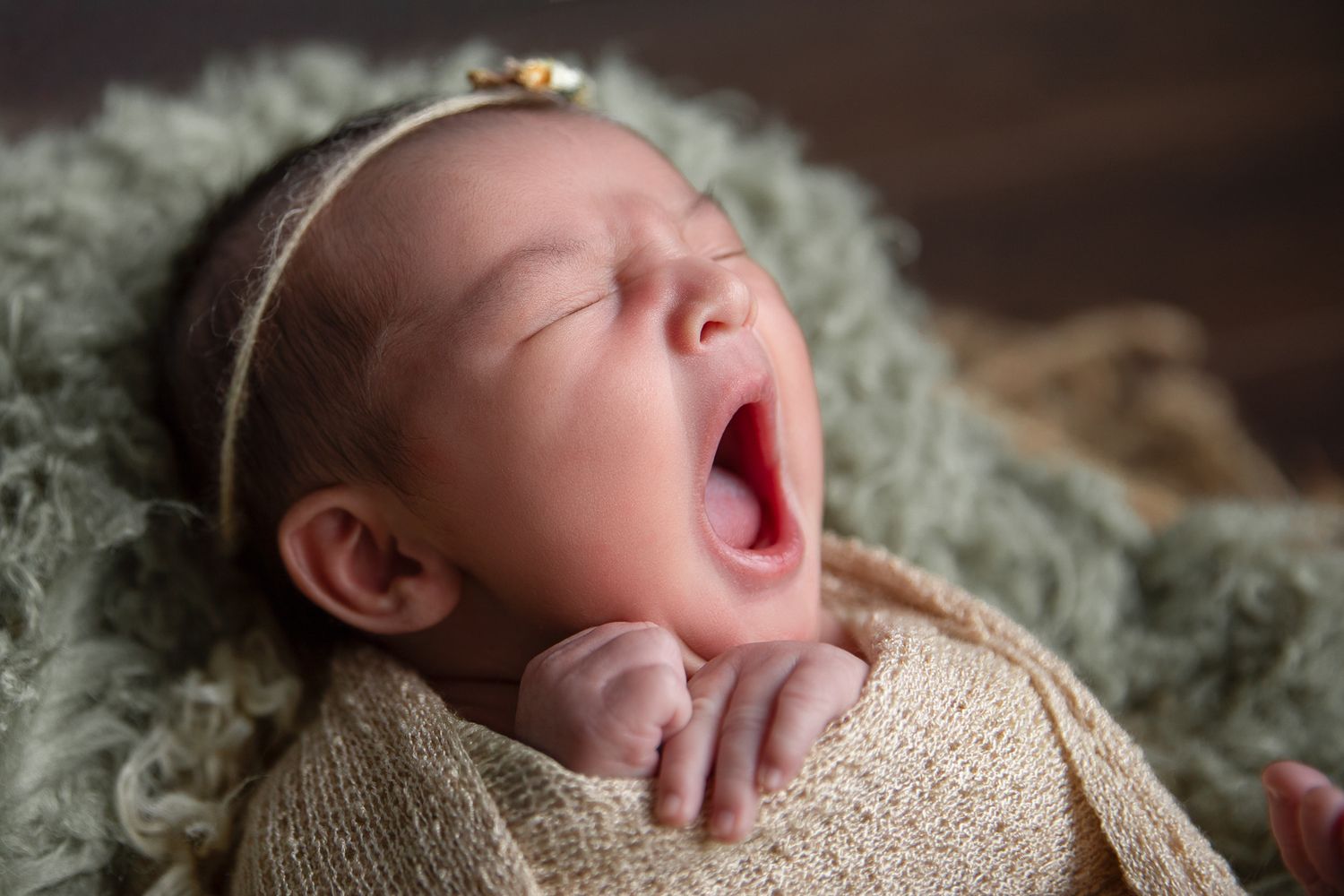 Newborn baby girl yawning while wrapped in a knit blanket, wearing a floral headband during a newborn photo session.