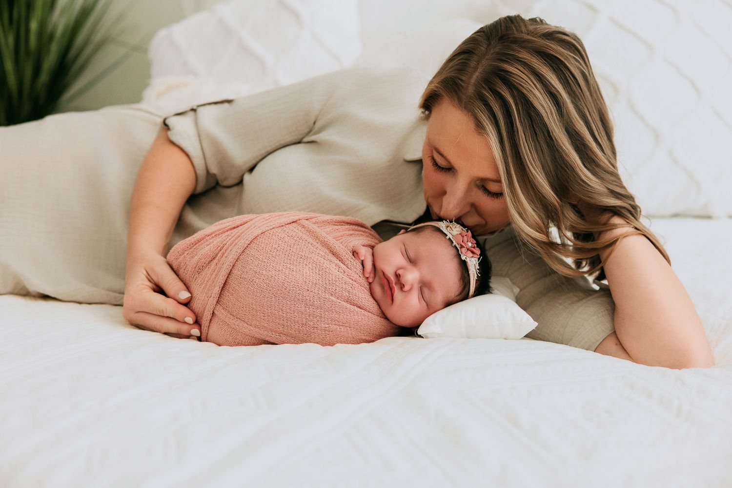 A mother tenderly kisses her swaddled newborn baby in a pink wrap on a white bed.