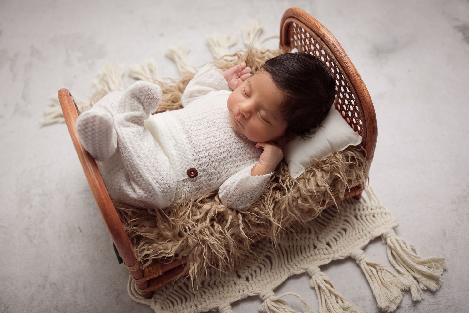 Newborn baby sleeping peacefully in a tiny wooden bed with cream knit outfit and fluffy blanket.