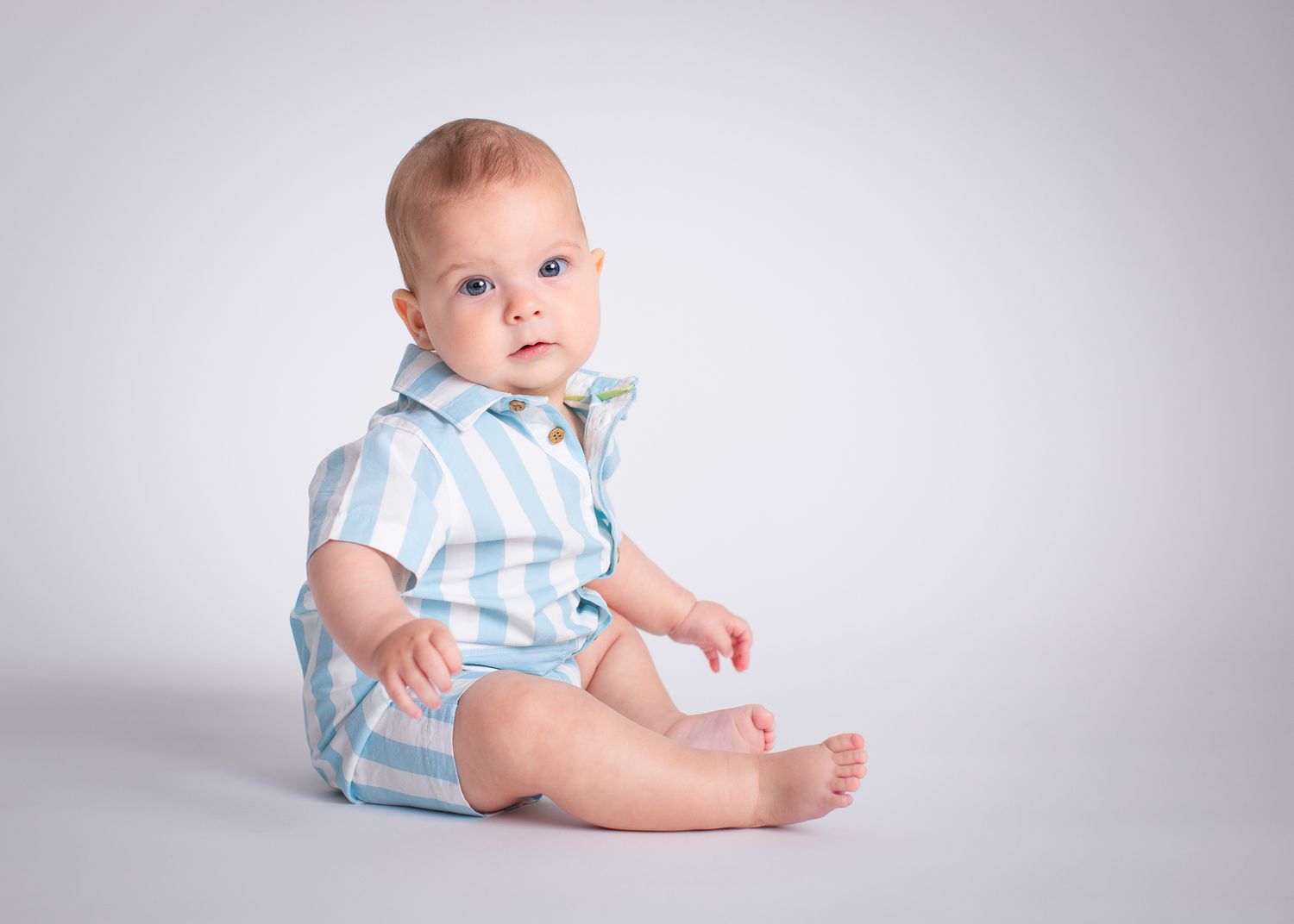 Adorable baby boy in blue striped onesie sitting on white studio background.