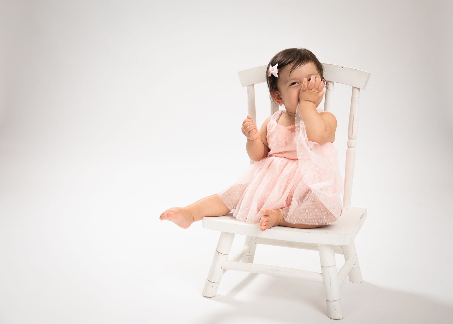 Toddler girl in pink tutu dress sitting on white chair covering her face.