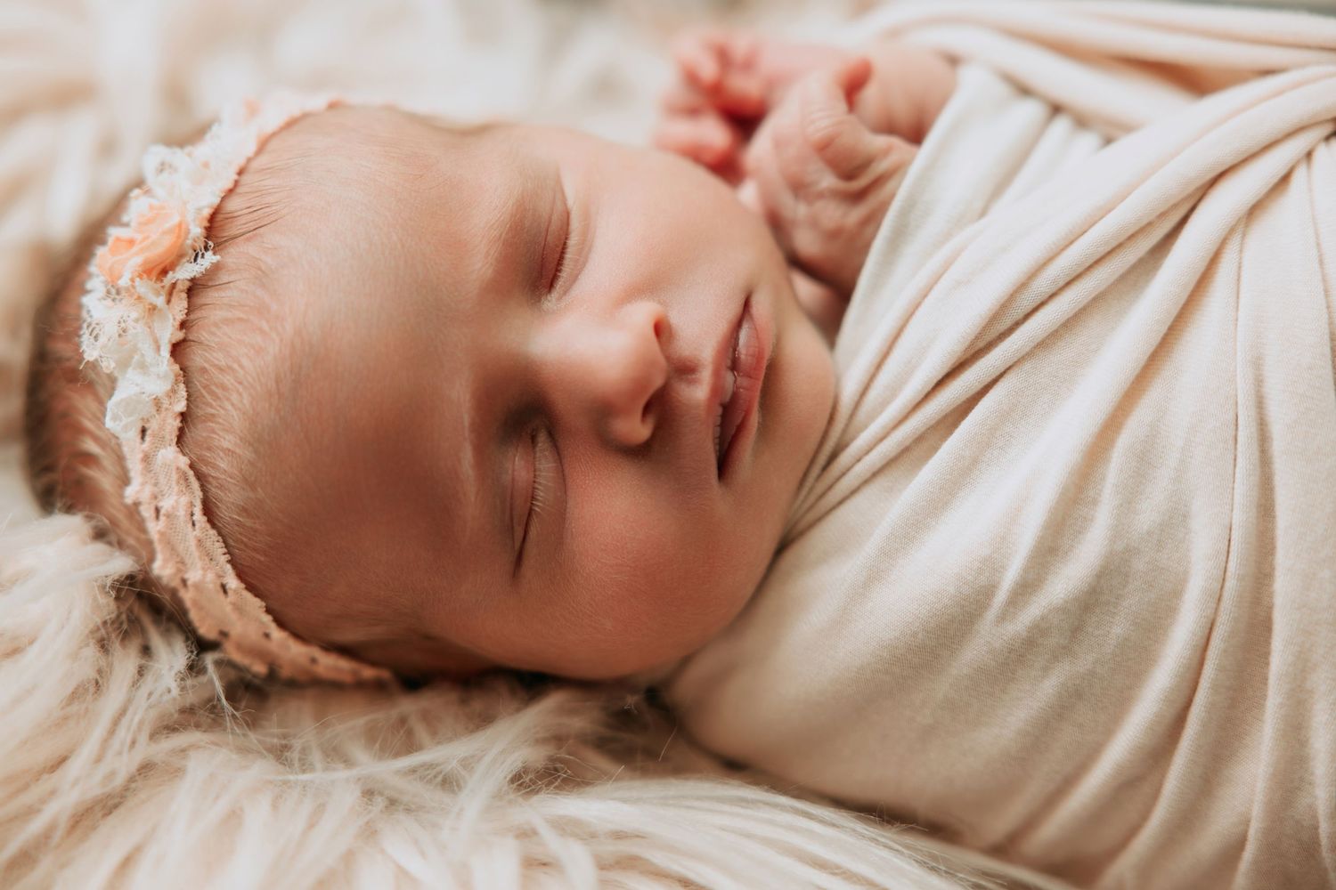 Peaceful newborn baby girl sleeping swaddled in cream wrap, wearing delicate floral headband, soft fur backdrop.
