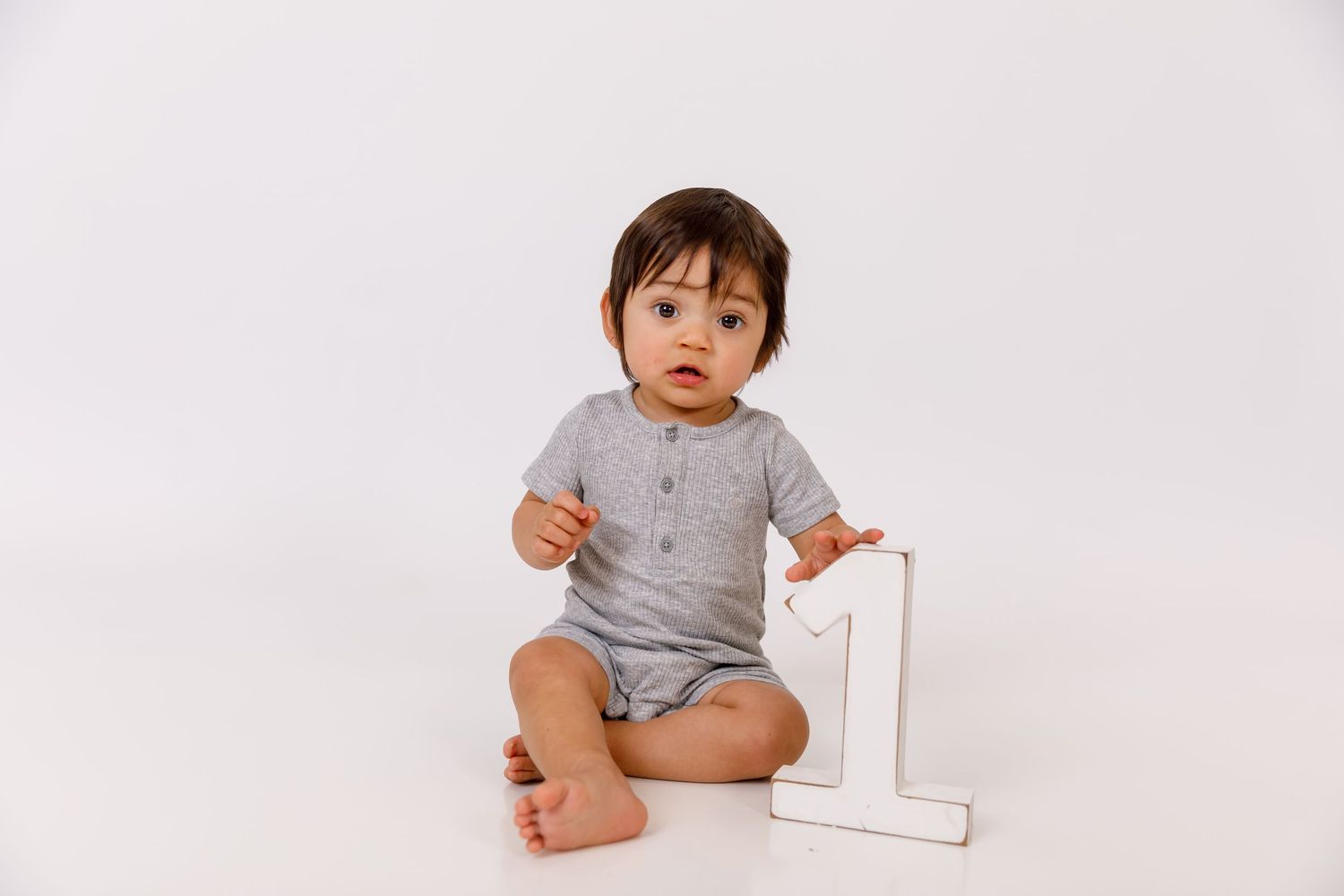 A baby in a gray onesie sits on a white floor next to a white number one, celebrating their first birthday.