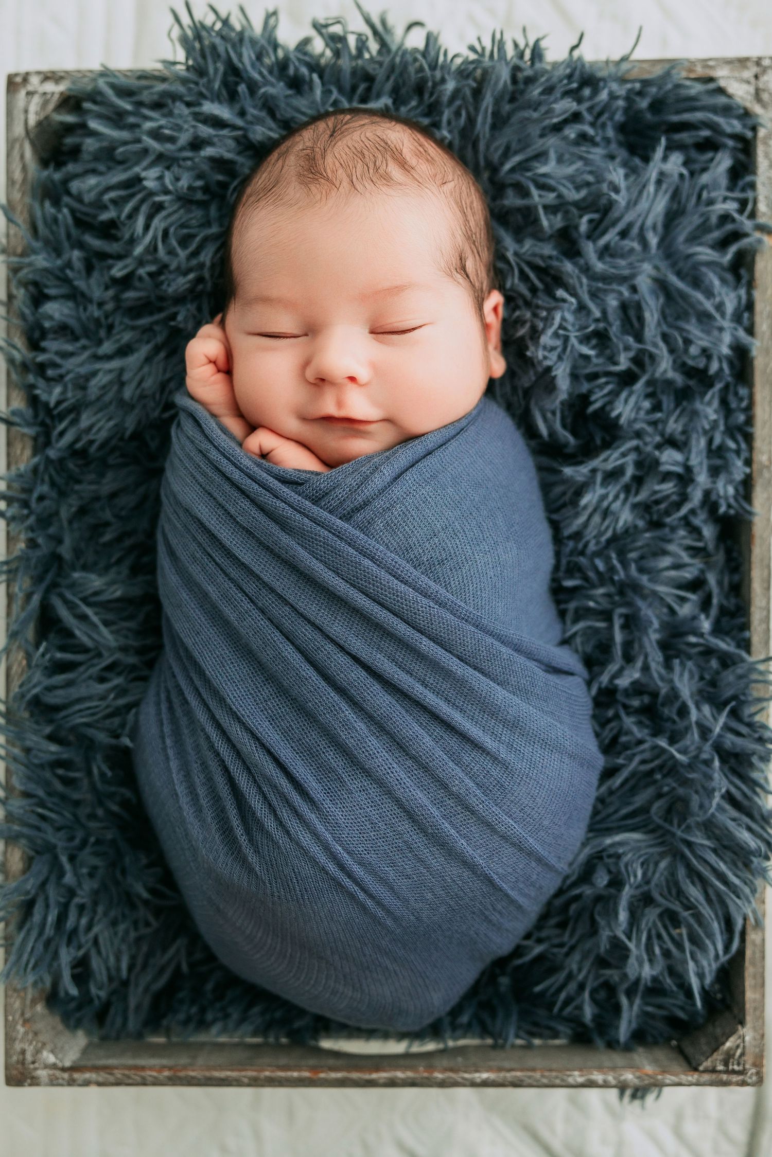 Peaceful newborn baby swaddled in blue wrap lying on fluffy blue rug in wooden crate.