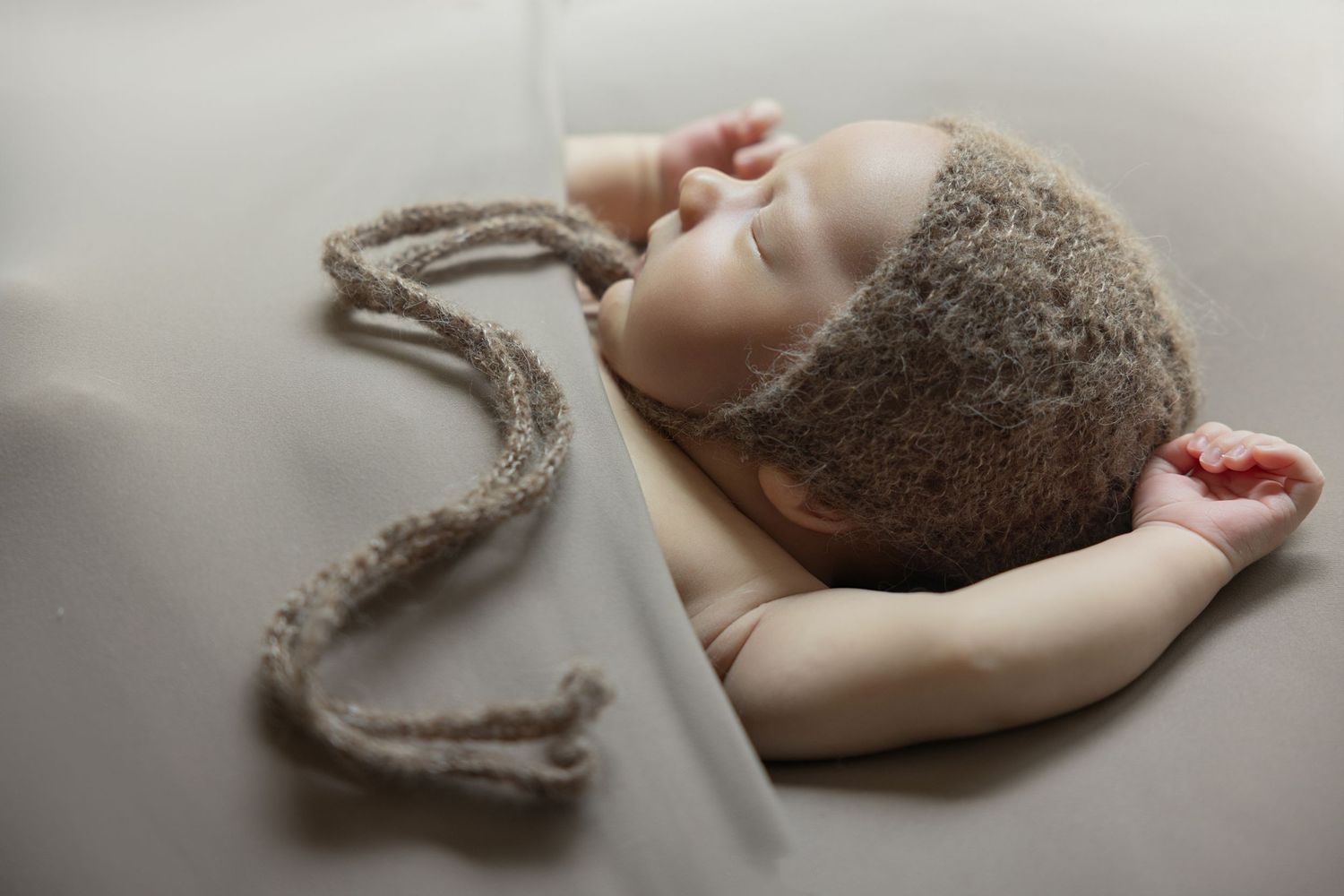 Peaceful newborn baby sleeping in a knit brown hat with long ties on a soft gray background.
