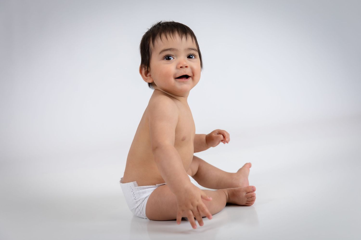 Smiling baby in a white diaper sitting on a white floor, looking up with a happy expression.