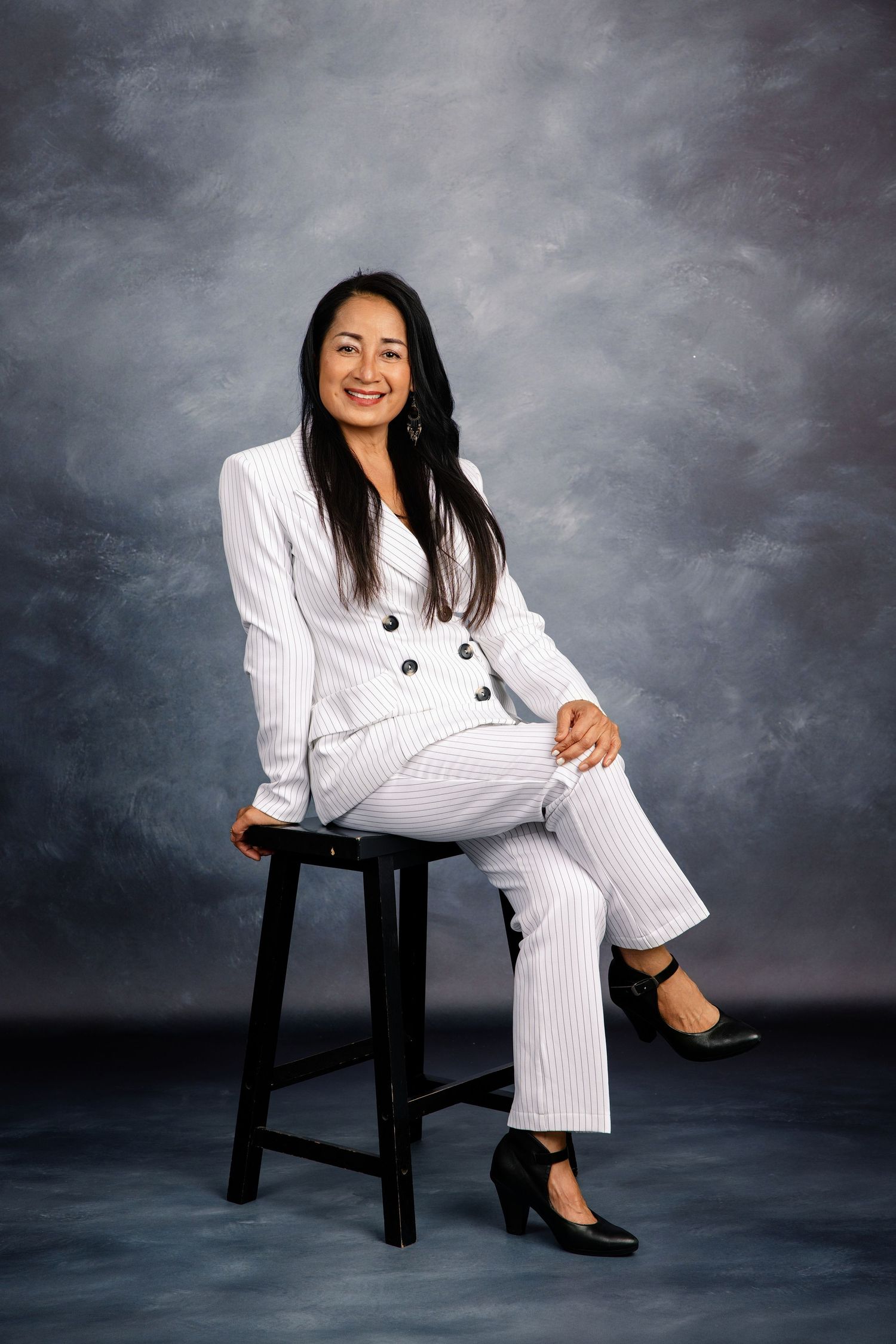 Woman in white pinstripe suit seated on stool, smiling confidently in professional studio portrait photo.